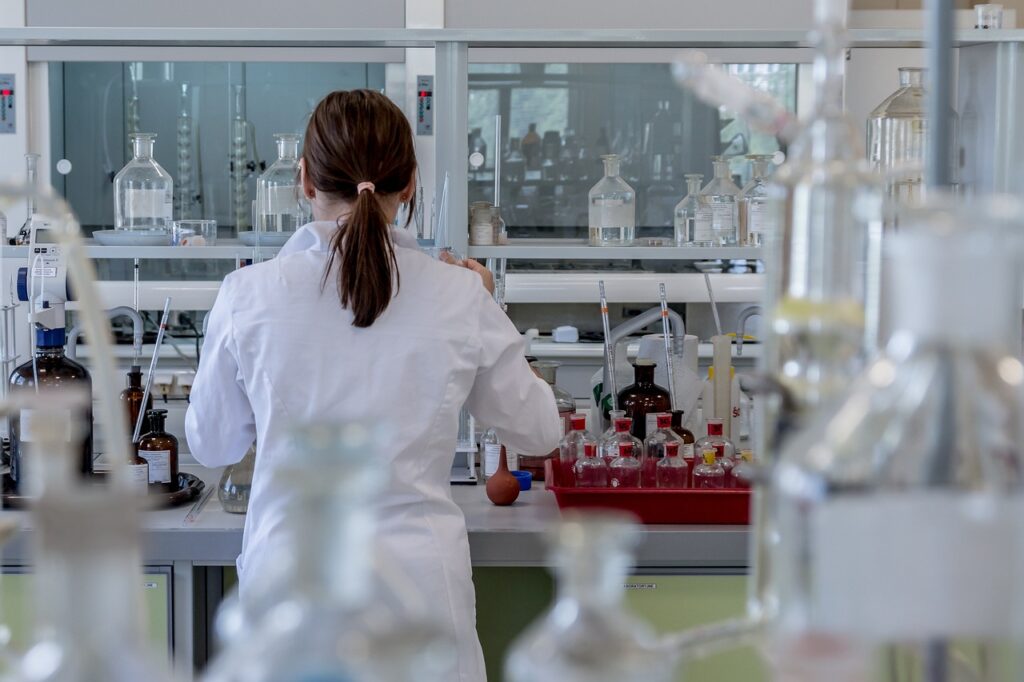 A female scientist in a white lab coat working in a modern laboratory surrounded by glassware and chemical bottles.