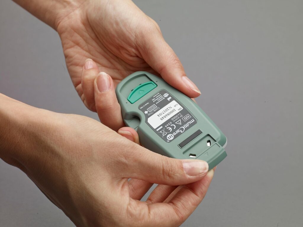 Hands holding a green handheld medical device, with visible regulatory labels and a removable compartment cover.