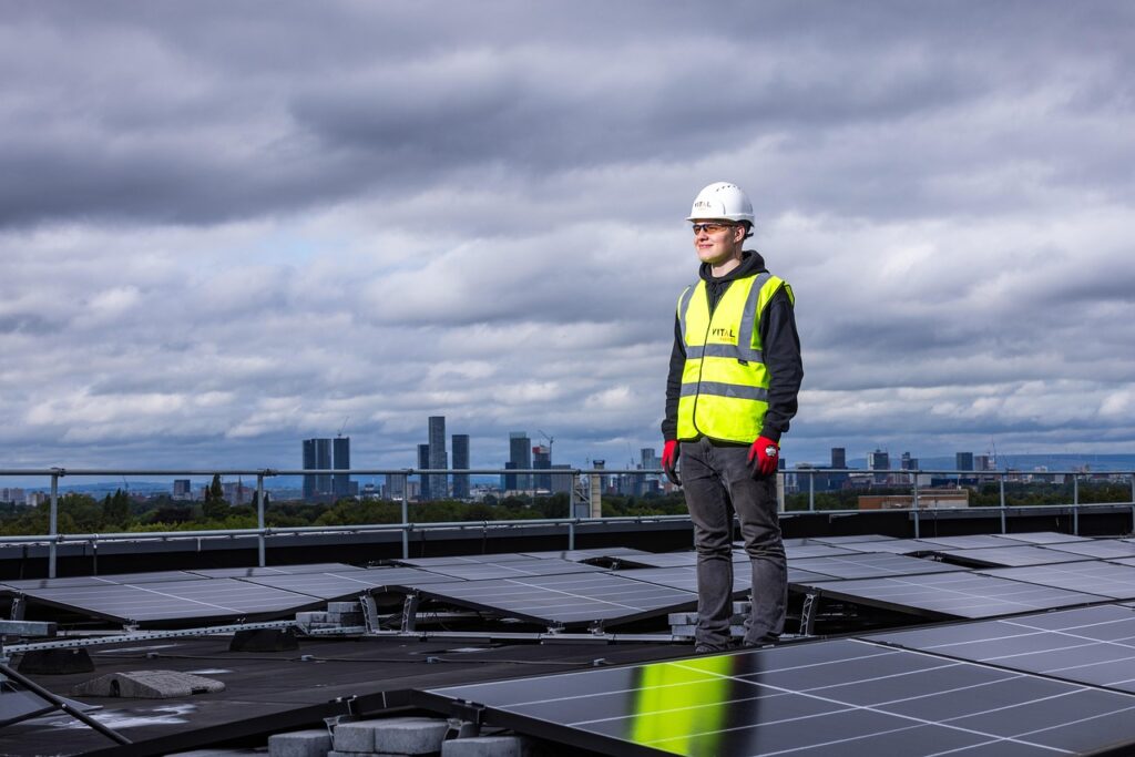 Worker wearing safety gear standing on a rooftop among solar panels, with a city skyline in the background under cloudy skies.