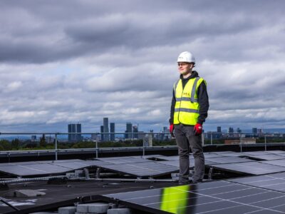 Worker wearing safety gear standing on a rooftop among solar panels, with a city skyline in the background under cloudy skies.