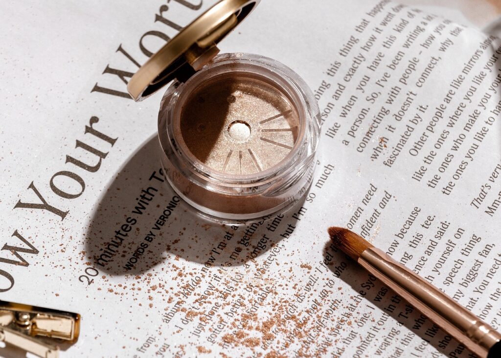 Open container of shimmery bronze mineral eyeshadow with a makeup brush and scattered powder on a magazine page.