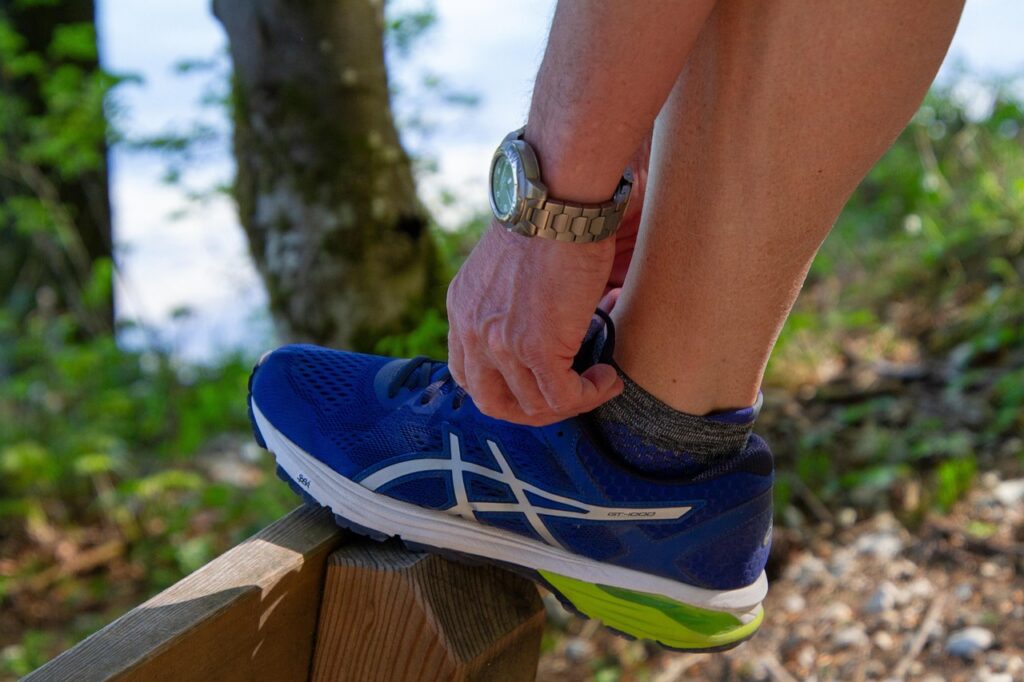 A person wearing a wristwatch bends down to tie the laces of a blue running shoe while resting their foot on a wooden fence outdoors.