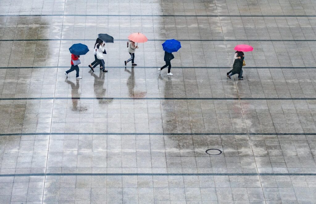 Six people walking on a wet tiled surface with umbrellas of different colors during a light rain.