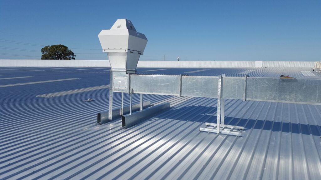 HVAC ventilation unit mounted on a metal rooftop with visible ductwork under a clear blue sky.