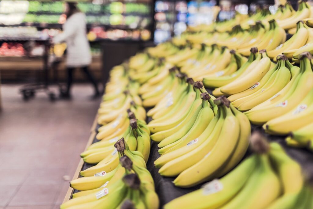 Display of ripe bananas arranged in rows at a grocery store, with a shopper pushing a cart in the background.