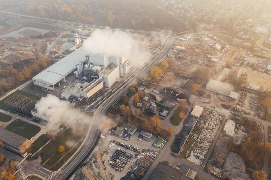 Aerial view of an industrial facility emitting steam or smoke, surrounded by construction materials, machinery, and forested areas in the background.