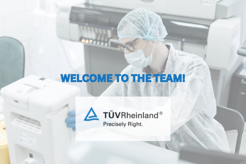 A male researcher tweaks the focus settings of a microscope sitting at a lab bench with test tubes. TUV Rheinland logo is in the foreground.