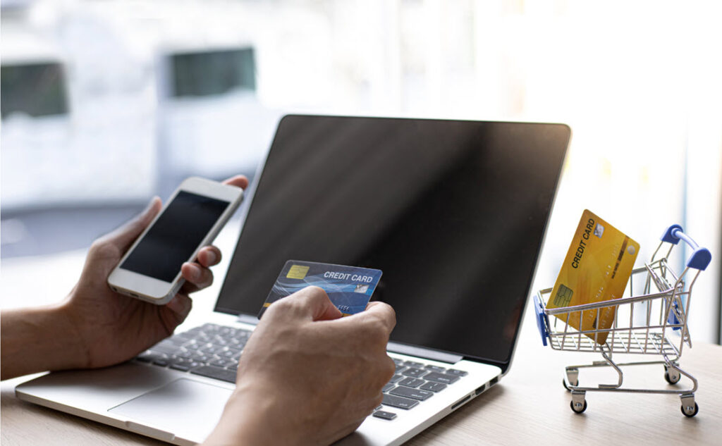 A person holding a smartphone in the left and credit card in the right hand sits in front of a laptop.