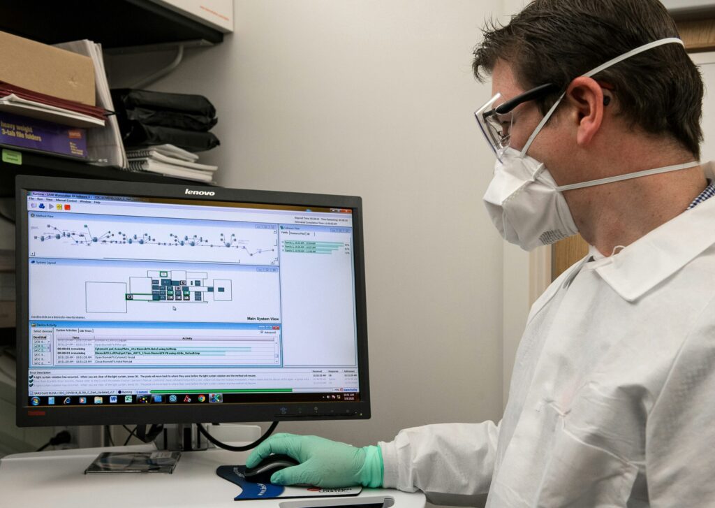 A scientist wearing PPE looks on at a computer showing scientific data.
