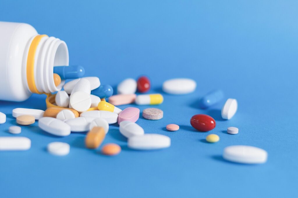 An open white bottle with various colorful pills and capsules spilling out onto a blue surface.