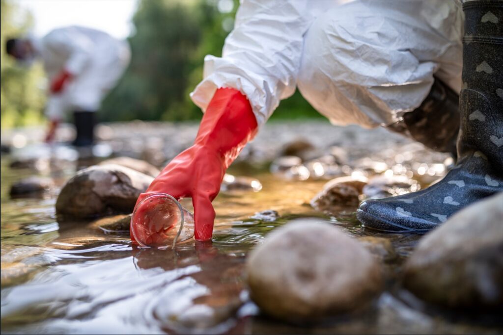 image of a scinetist in protective clothing and gloves collecting a water sample for environmental microbiology testing