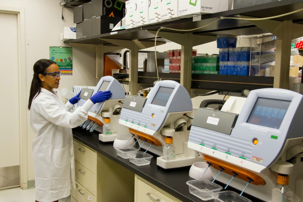 A female researcher in lab coat and gloves examines a series of laboratory instruments.