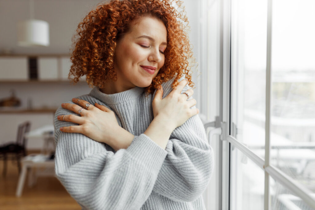 a red heded woman standing next wo a window caressing a grey sweater suggesting the importance of surface and texture testing in the consumer goods industry