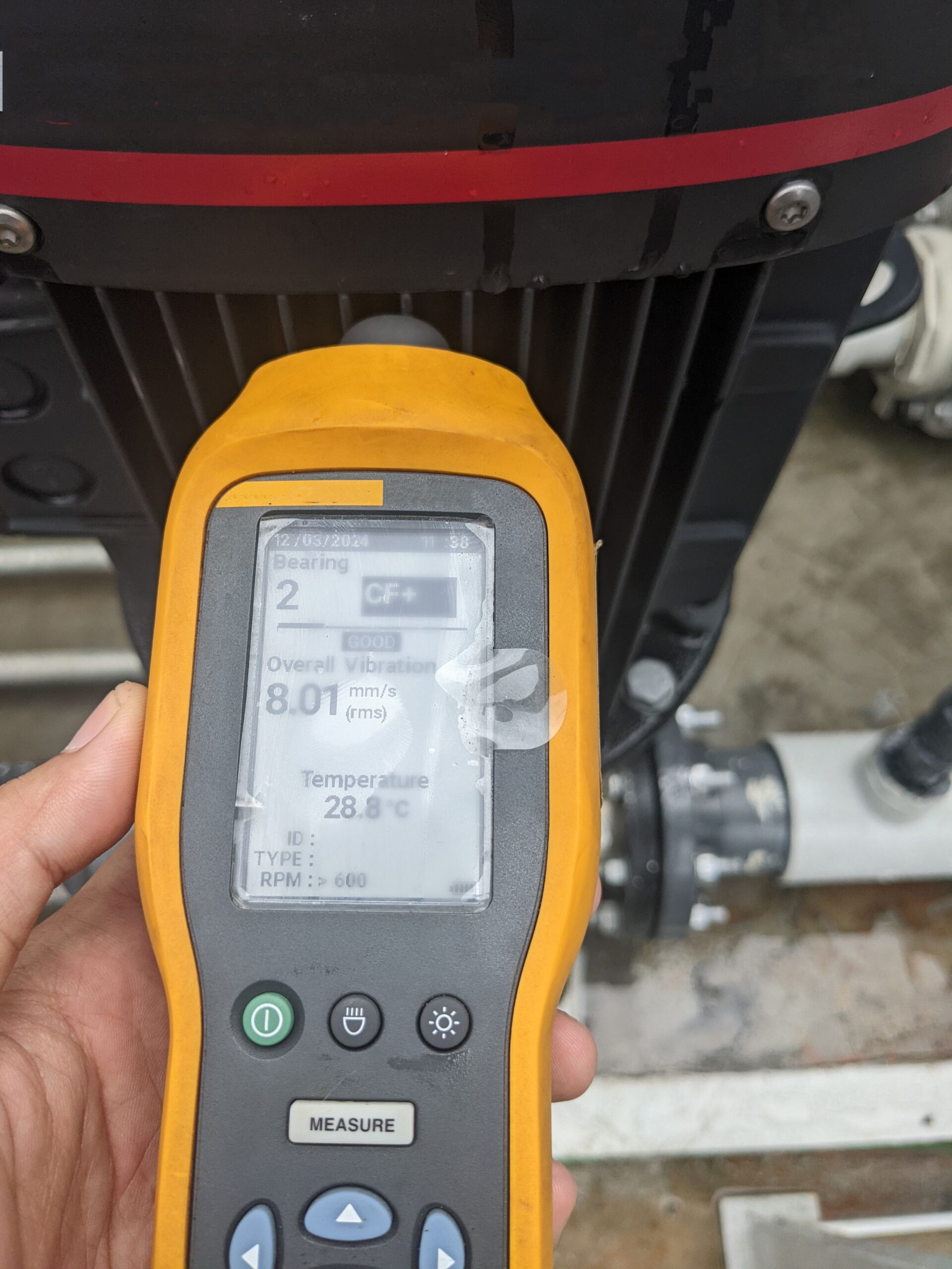 A close-up shot of a Fluke vibration meter displaying measurements. The meter is held in a hand and is being used to measure the vibration levels of a machine.