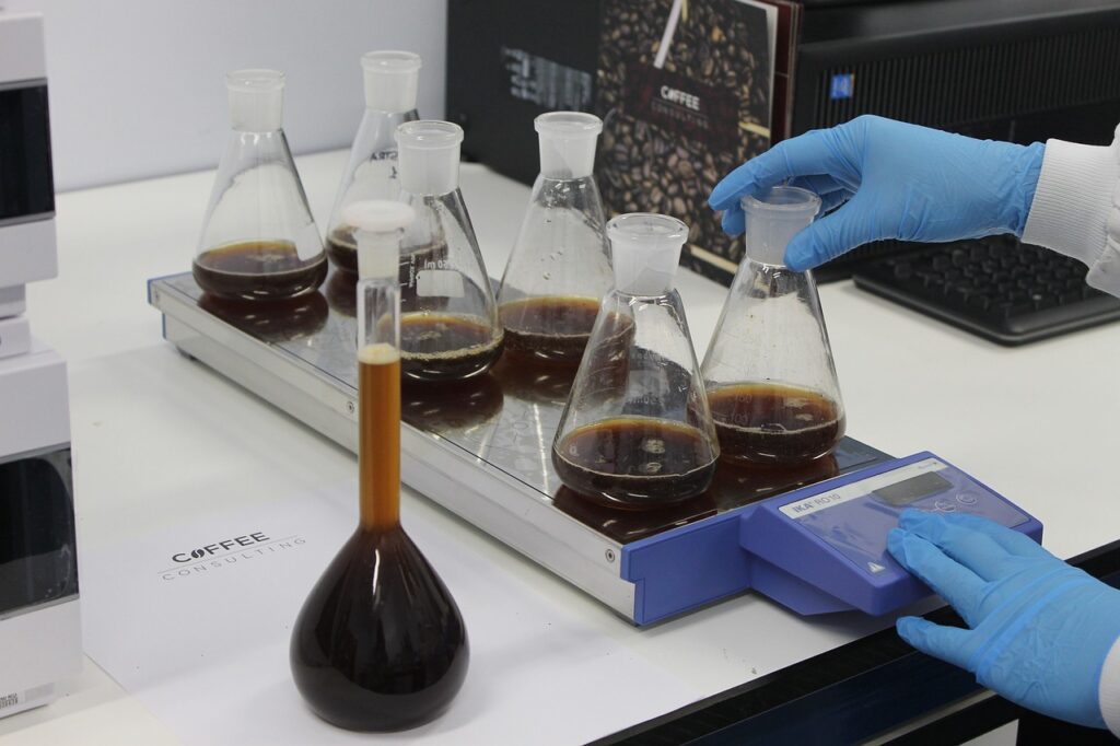 Gloved hands operating lab equipment with multiple Erlenmeyer flasks filled with dark liquid, likely coffee samples, on a lab bench.