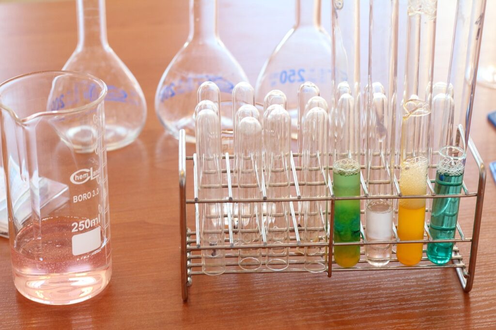 Glass test tubes in a metal rack, some filled with colorful liquids, alongside laboratory flasks and a beaker on a wooden surface.