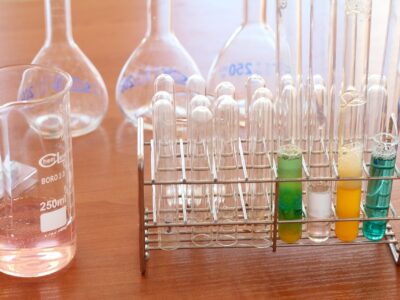 Glass test tubes in a metal rack, some filled with colorful liquids, alongside laboratory flasks and a beaker on a wooden surface.