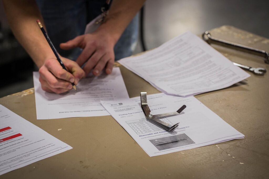 Person filling out paperwork at a workbench with technical documents, inspection tools, and a wrench nearby.