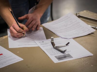 Person filling out paperwork at a workbench with technical documents, inspection tools, and a wrench nearby.